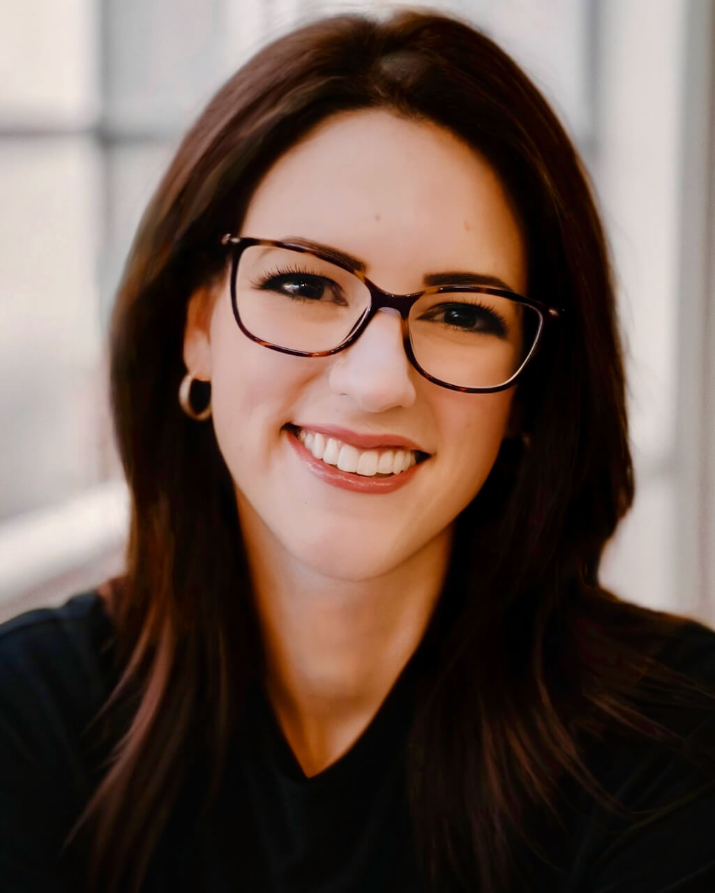 A woman with shoulder-length dark brown hair and glasses smiles warmly at the camera. She is wearing small hoop earrings and a black top, with a softly blurred background suggesting an indoor setting with natural light.