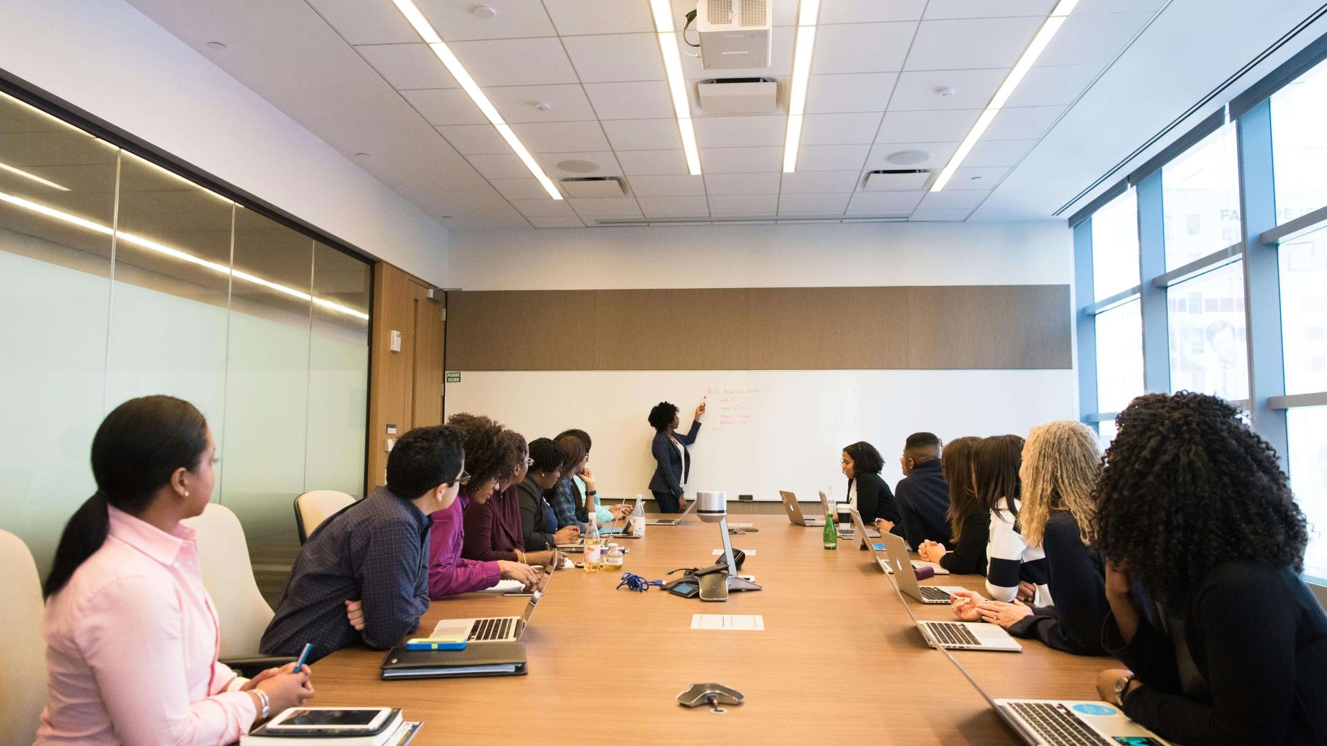A diverse group of professionals sits around a long conference table with laptops open while one person stands at a whiteboard leading a collaborative discussion in a modern meeting room.