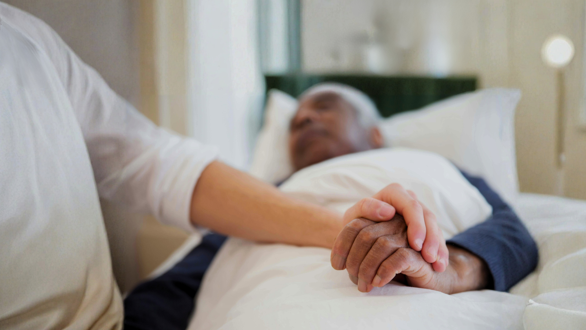 Older adult holding caregiver's hand in bed