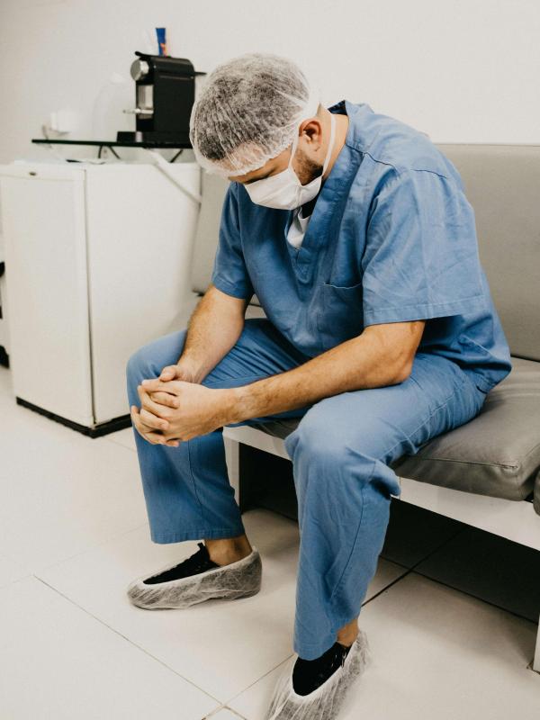 A healthcare professional in scrubs sits on a bench in a clinical room with head bowed and hands clasped, appearing emotionally distressed or exhausted after a difficult moment at work.