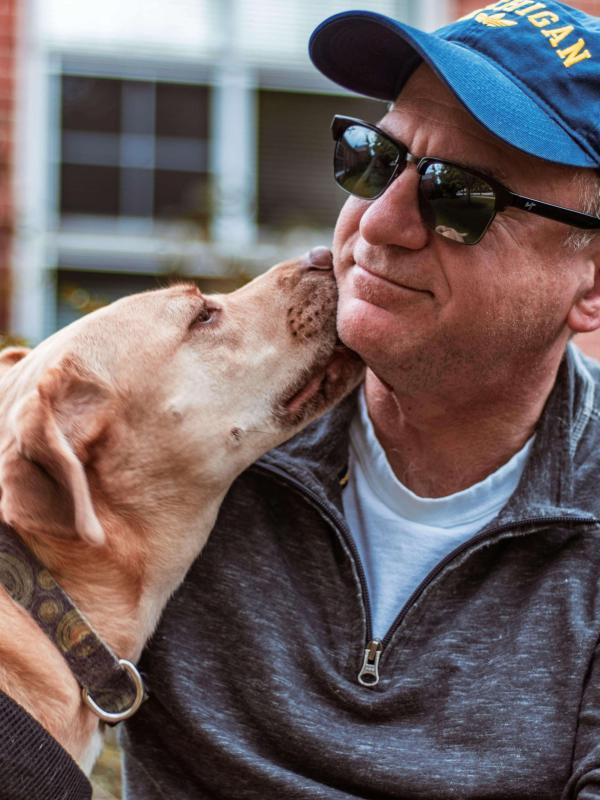 Person sitting outdoors wearing a hat and sunglasses, holding a dog that is looking up at them affectionately.