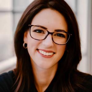 A woman with shoulder-length dark brown hair and glasses smiles warmly at the camera. She is wearing small hoop earrings and a black top, with a softly blurred background suggesting an indoor setting with natural light.