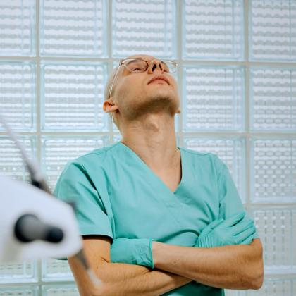 A healthcare professional in scrubs leans back slightly with arms crossed and head tilted upward, appearing to take a brief pause or stretch in a clinical setting.